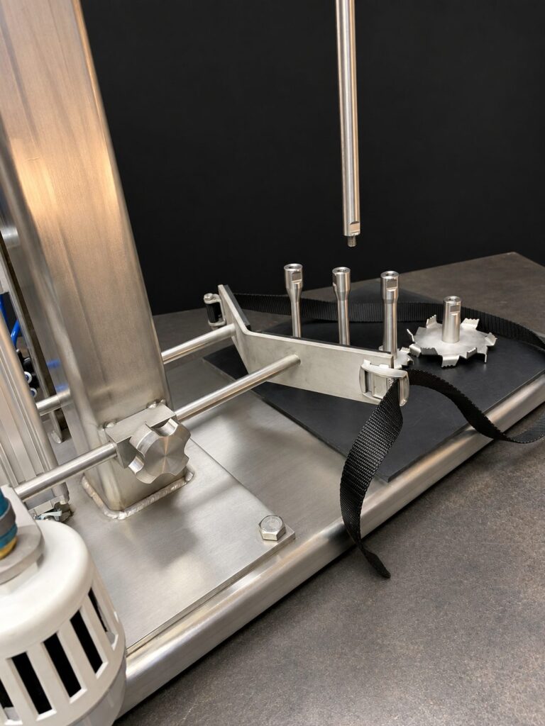 Stainless benchtop mixer base plate with a rubber pad and strap, an adjustable container yoke locked by a knob clamp, a threaded shaft end above, and interchangeable quick-change blade stubs.