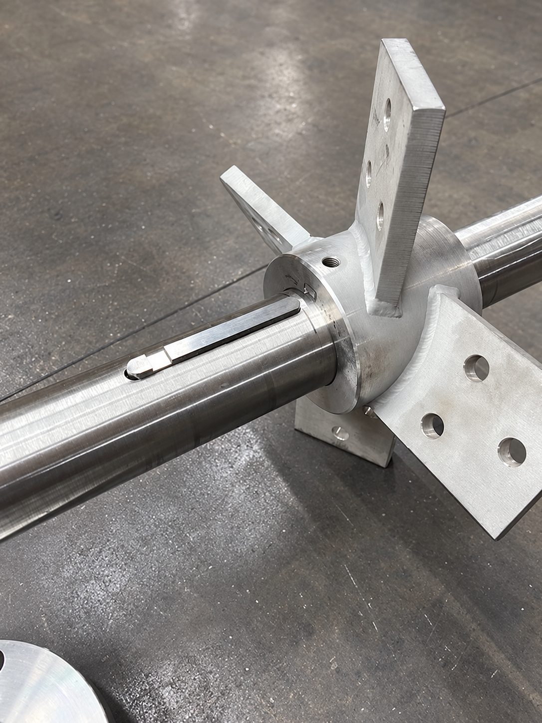 A machined impeller hub welded to a shaft with four angled blade mounting plates, lying on a shop floor with the shaft keyway and key visible.