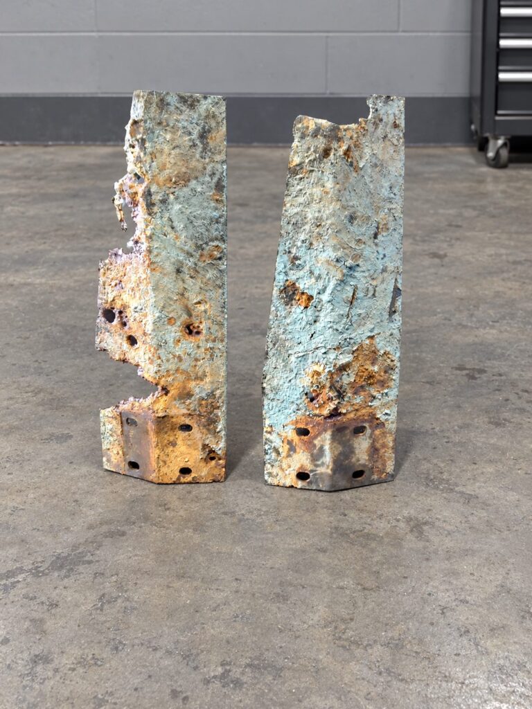 Two heavily pitted curved steel sections with flaking paint and bolt holes at one end, standing on a shop floor.