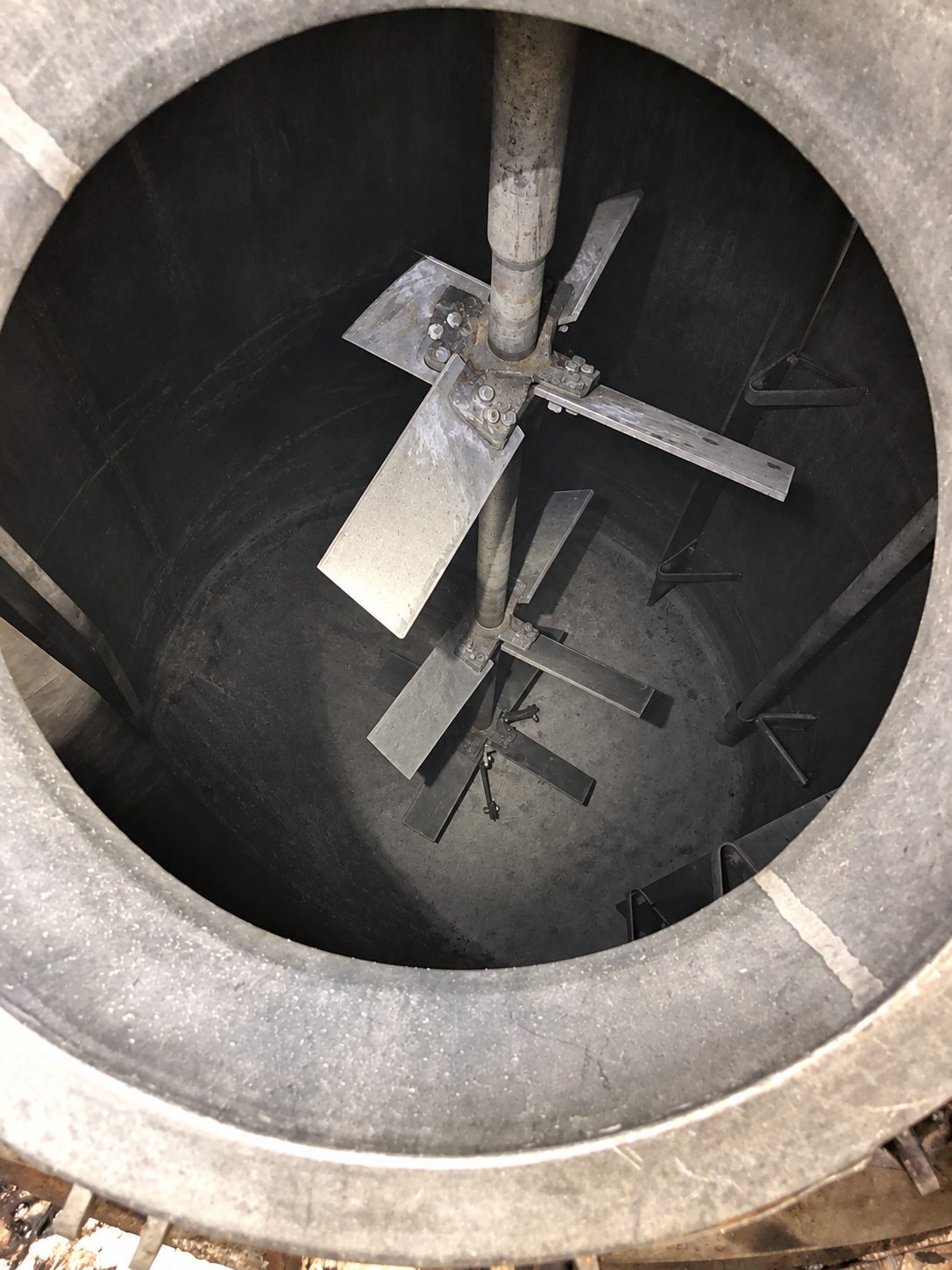 View down through a tank opening onto a mixer shaft fitted with two bolted pitched blade turbines, with flat wall baffles bracketed to the vessel wall around them.