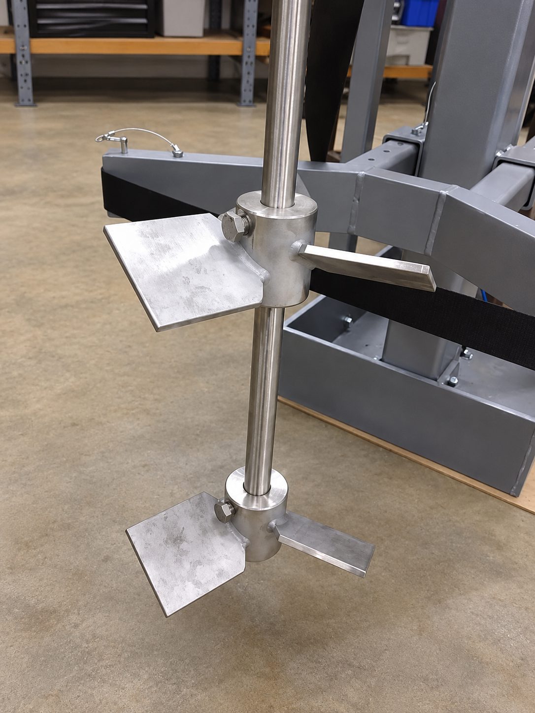 Stainless mixer shaft carrying two pitched blade impellers, each with flat blades bolted to a split clamp hub, hanging above the painted base frame of a stand.